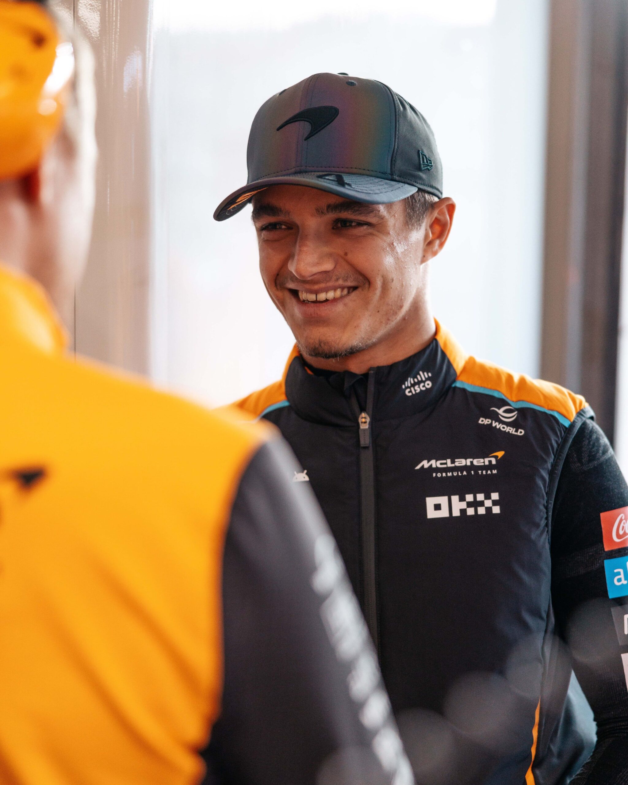 Lando Norris in a McLaren suit, looking over a McLaren employee