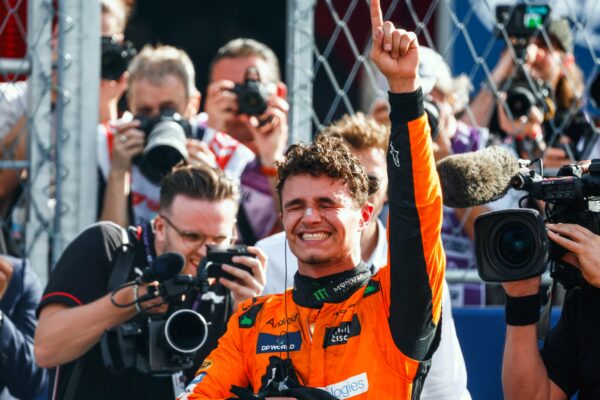 Lando Norris pointing his index finger in the air with his eyes closed. Photographers in the background